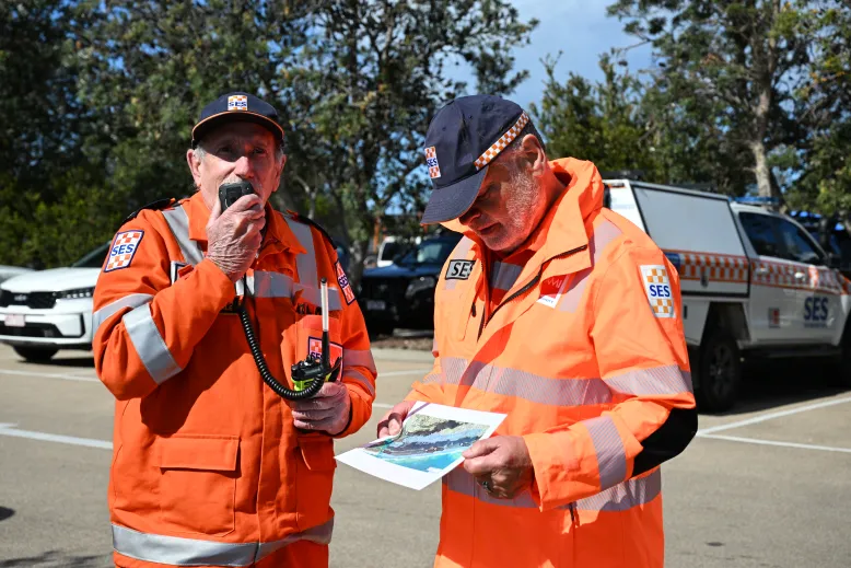 Two men in orange VicSES uniforms and navy blue caps stand next to each other. One man is holding a radio microphone and the other is looking at a map.