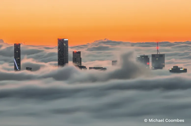 City towers rise above a sweeping layer of opaque low cloud beneath a glowing orange twilight sky.