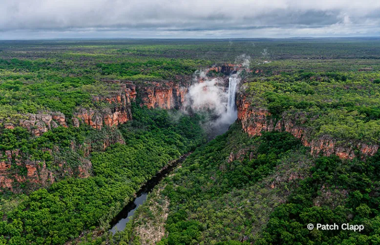 Waterfall plunges into a mist-filled gorge surrounded by dense forest and towering red cliffs.