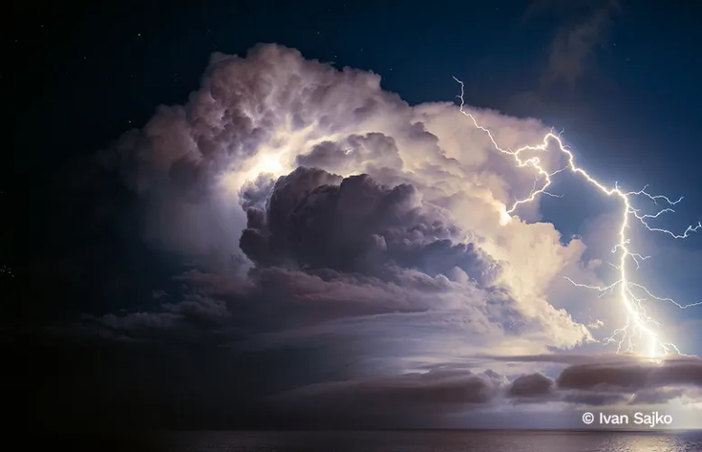 Forked lightning illuminates a towering thunderstorm cloud above a dark and tranquil sea.