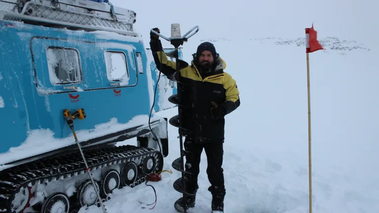 Man in waterproof gear stands in front of blue vehicle with caterppillar tread in Antarctica's icy landscape. He holds a spiral piece of equipment used to drill sea ice.