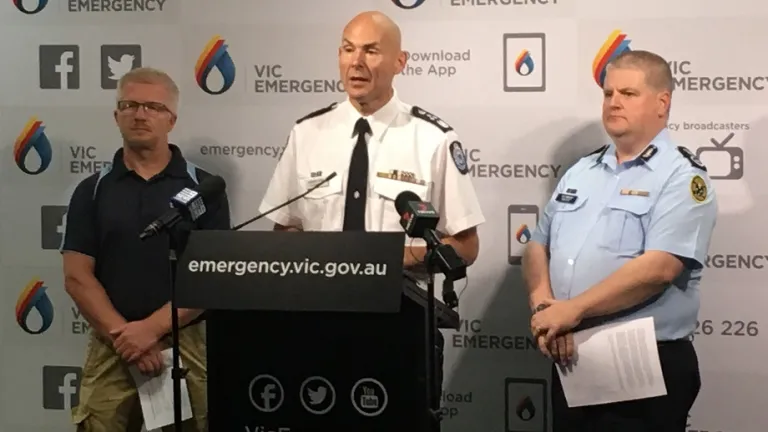 Press conference with representatives from the Bureau of Meteorology, Emergency Management Commissioner and State Emergency Services standing in front of a wall with Vic Emergency logos. The Commissioner is in the centre, speaking into a microphone at a podium that carries the Vic Emergency contact details – emergency.vic.gov.au, 1800 226 226, and social media logos for Twitter, Facebook and YouTube. The Bureau and SES representatives are standing at either side holding speaking notes.