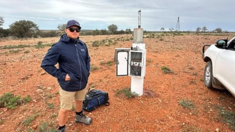 Field technician Darcy stands on a red dirt plain under a cloudy sky, with a bag at his feet. Nearby, observing equipment has the service door open. A vehicle is partly visible on the right of the image.