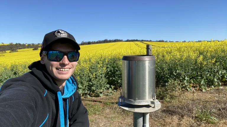Field technician Kyle stands next to a rain gauge, wearing sunglasses and a cap. Behind him, a bright yellow canola field stretches to the horizon under a clear blue sky.