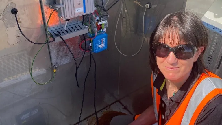 Person in an orange high-visibility vest kneels inside a metal equipment cabinet, inspecting wiring and battery components attached to an electronic control unit.